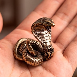Op de afbeelding: Een klein, metallic bronskleurig cobra-beeldje met een opgerold lichaam en een rechtopstaande, gespreide kap. De slang is afgebeeld met een open bek, waarbij de giftanden zichtbaar zijn, en heeft gedetailleerde, textuurrijke schubben.