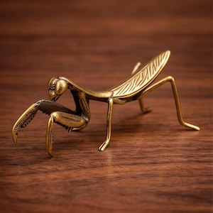Könnte beinhalten: Eine detaillierte Messing-Gottesanbeterin-Figur. Das Insekt ist auf einer Holzoberfläche platziert und zeigt sein aufwendiges Design. Die Beine und der Körper der Figur sind präzise gefertigt und heben die einzigartigen Merkmale des Insekts hervor.