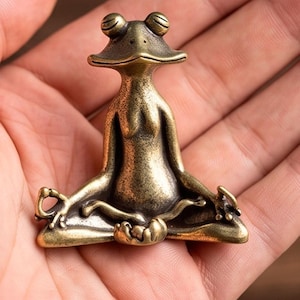 Puede incluir: Una pequeña figura de latón de estilo antiguo que representa una rana sentada en posición de meditación con las piernas cruzadas. La rana tiene un acabado metálico liso con zonas oscurecidas, ojos prominentes y manos en gesto de mudra.