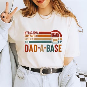 May include: A cream-coloured t-shirt featuring the text "MY DAD JOKES STAY SAFELY SAVED IN THE DAD-A-BASE" in a retro style. The design incorporates a floppy disk icon and horizontal stripes in red, orange, brown, teal, and blue.