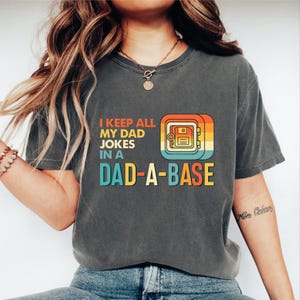 Op de afbeelding: Donkergrijs t-shirt met de tekst "I KEEP ALL MY DAD JOKES IN A DAD-A-BASE" in oranje, blauwgroen en geel. De afbeelding toont een retro-kluis met een diskette-ontwerp.