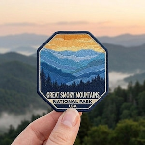 May include: An octagonal embroidered patch featuring a landscape of the Great Smoky Mountains National Park. The design displays layers of blue mountains, a dark forest silhouette, and a yellow sky, with the text GREAT SMOKY MOUNTAINS NATIONAL PARK USA.