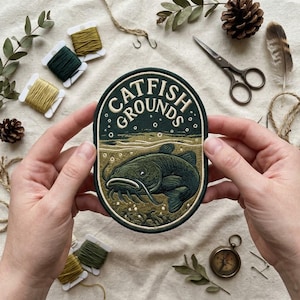 May include: An oval embroidered patch featuring a dark green background with a catfish swimming underwater. The text CATFISH GROUNDS is stitched in cream-colored thread at the top. The design includes bubbles and water ripples in shades of green and cream.
