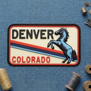 May include: A rectangular patch featuring the text "DENVER" above "COLORADO" in black and red, respectively. A blue and black rearing horse is centered on a cream background, with red and blue stripes. The patch has a black border.