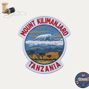 May include: A circular embroidered patch featuring Mount Kilimanjaro with a snow-capped peak, clouds, and trees in a landscape. The text MOUNT KILIMANJARO and TANZANIA is stitched in red. Includes two spools of thread and a small circular logo.