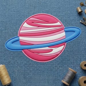 May include: A pink and white planet-shaped patch with a blue ring. The patch has a layered design with a pink outline. The background is a blue fabric, with spools of thread and sewing notions scattered around the patch.