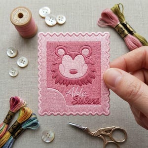 May include: A rectangular pink embroidered patch with a scalloped edge featuring a stylized bear face and the text "Able Sisters" in cursive script. The patch is surrounded by sewing supplies including thread spools, buttons, and scissors.