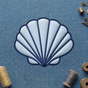 May include: A scallop shell patch featuring a light blue center with raised ridges and a dark blue border. The patch is centered on a textured blue fabric background, surrounded by sewing supplies including thread spools and a metal thimble.