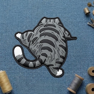 May include: A gray and black cat-shaped patch, viewed from the rear, with striped markings. The patch has a white paw and tail tip. The patch is on a blue fabric background, with sewing notions in the background.