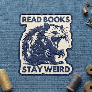 May include: A blue and white patch with the text "READ BOOKS STAY WEIRD" and a detailed illustration of an opossum with its mouth open. The patch is on a blue fabric background, with spools of thread and sewing tools.