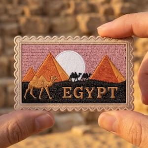 May include: A rectangular embroidered patch with a scalloped border featuring a desert scene with orange pyramids, a white sun, and black silhouettes of camels against a pink sky. The word EGYPT is stitched in gold thread on a black base.