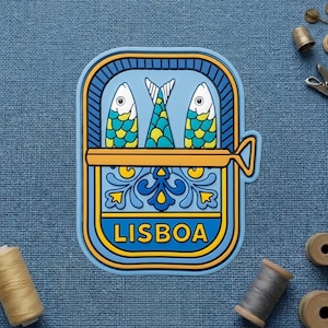 May include: A colorful Lisbon-themed magnet featuring a sardine can design. The magnet has a blue background with a yellow border and the word "LISBOA" in white. The can contains three stylized fish with yellow and blue scales.
