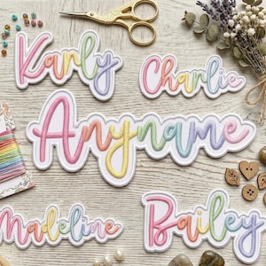 May include: Custom embroidered name patches featuring a colorful pastel gradient script font. The set includes individual patches with the names Karly, Charlie, Anyname, Madeline, and Bailey, each with a white border on a light wood surface.