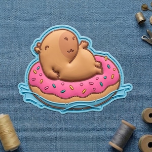 May include: A whimsical patch featuring a cartoon character lounging on a pink donut float with colorful sprinkles. The character is light brown, and the donut has a light brown base with a blue outline. A fun, playful design.