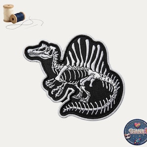 May include: Black and white embroidered patch featuring a Spinosaurus dinosaur skeleton. The patch has a detailed design with a white outline and is set against a black background. Includes spools of thread and a needle.