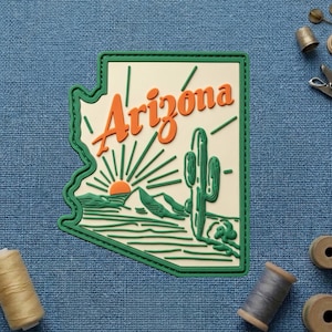 May include: A rectangular patch featuring the outline of Arizona in green, with the word "Arizona" in orange script. The design includes a cactus, mountains, and a setting sun. The patch is on a blue textured surface.