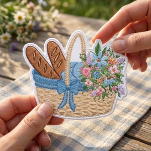 May include: An embroidered patch featuring a tan woven basket filled with two brown baguettes and a bouquet of blue, pink, and purple flowers. The basket is decorated with a blue ribbon tied in a bow at the center, all outlined in white.