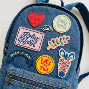May include: A denim backpack adorned with colorful patches. Patches include a red heart with "hug it out", a pink rectangle with "Relax Hard", a yellow circle with "Let's do this", and a blue banner that says "Dream in Color".