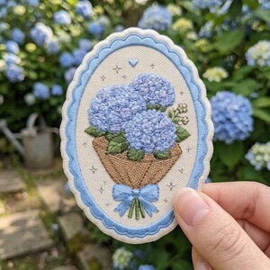 May include: An oval-shaped embroidered patch featuring a bouquet of blue hydrangea flowers wrapped in brown paper with a light blue ribbon bow. The design is set against a cream background with small star and heart accents, framed by a scalloped blue border.
