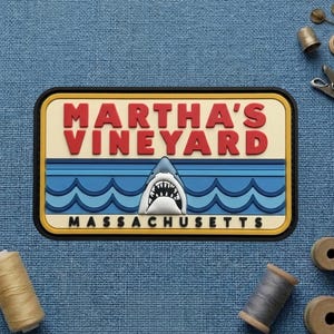 May include: A rectangular patch with the words "MARTHA'S VINEYARD" in red block letters. Below, a shark emerges from blue waves. The text "MASSACHUSETTS" is at the bottom. The patch has a black border.