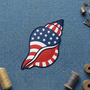 May include: A rubberized patch shaped like a seashell featuring a patriotic design with a dark blue section containing white stars and alternating red and white stripes across the spiral shell body, presented on a textured blue fabric background.