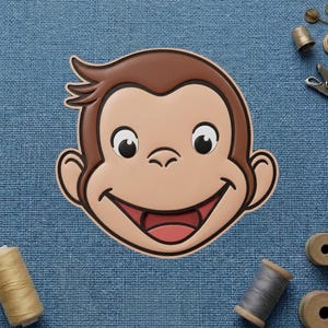 May include: A smiling monkey face with brown hair and a light brown face, outlined in black. The monkey's mouth is open, revealing a pink tongue. The background is a blue fabric with sewing supplies.