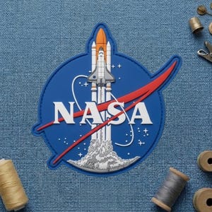 May include: A blue, white, red, and orange NASA patch featuring a space shuttle launching. The patch has the NASA logo and a red swoosh. The patch is on a blue textured background.