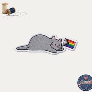May include: An embroidered patch featuring a gray cat lying down and holding a small rectangular flag with rainbow and progress pride stripes. The patch has a white border. Two wooden spools of thread and a needle with black thread are visible.