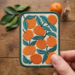May include: A rectangular embroidered patch featuring a repeating pattern of bright orange citrus fruits with dark teal green leaves on a cream-colored background, finished with a dark teal green stitched border around the entire edge.