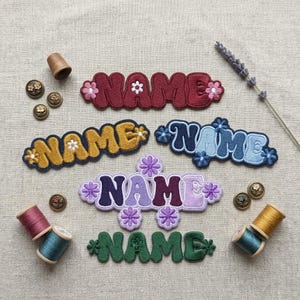 May include: Five embroidered patches featuring the word NAME in various colors including burgundy, gold, blue, purple, and green. Each patch is decorated with small flower motifs. Four spools of thread and several small metal buttons are included.