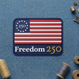 May include: A rectangular patch featuring a stylized American flag design with red and white stripes, a blue field with stars, and the text "Freedom 250". The patch is on a blue fabric background.