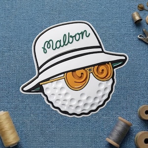 May include: A white golf ball character wearing a white hat with the word "Malbon" in green script, and orange sunglasses. The golf ball has a dimpled surface and is set against a blue background.