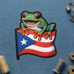 May include: A green frog with red eyes and orange feet is perched on a Puerto Rican flag patch. The flag features a white star on a blue triangle, with red and white stripes. The patch has a black border.
