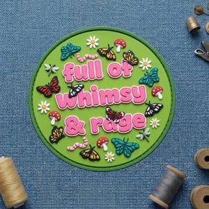 May include: A round, green decorative item with the words "full of whimsy & rage" in pink. The item is decorated with colorful butterflies, mushrooms, flowers, and a worm.