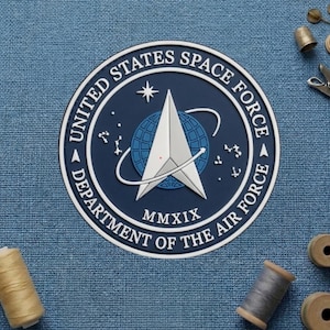 May include: A circular patch featuring the United States Space Force emblem. The design includes a white and silver delta symbol over a blue globe, surrounded by text that reads "UNITED STATES SPACE FORCE" and "DEPARTMENT OF THE AIR FORCE" in white. The background is a textured blue fabric.