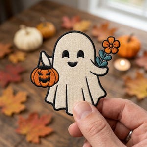 May include: An embroidered patch featuring a cream-colored ghost with a smiling face. The ghost holds a small orange jack-o'-lantern bucket in one hand and a teal-stemmed orange flower in the other. The design is outlined in black thread.