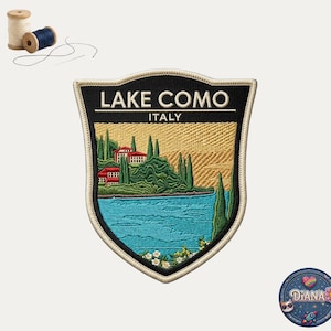 May include: A shield-shaped embroidered patch featuring the text LAKE COMO ITALY in white. The design depicts a blue lake, green cypress trees, and red-roofed buildings against a golden sky. Two wooden spools of thread are positioned above.