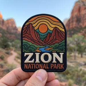 May include: An embroidered patch featuring a landscape of red rock mountains, a blue river, and a yellow sun against an orange sky. The text ZION NATIONAL PARK is stitched in white and orange lettering on a black background at the bottom.