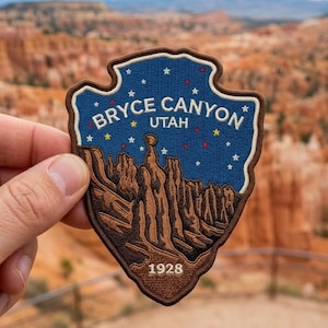 May include: A shield-shaped embroidered patch featuring a blue night sky with colorful stars above brown rock formations. The text reads BRYCE CANYON UTAH and 1928. The patch has a dark brown border and is shaped like an arrowhead or park emblem.