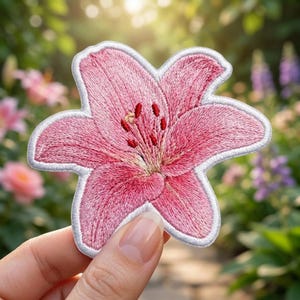 May include: An embroidered patch shaped like a pink lily flower with six petals, featuring detailed thread work in shades of light and dark pink, a white border, and small red stamens at the center of the bloom, showcasing intricate floral textures.