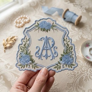 May include: A cream-colored fabric patch with a scalloped edge, featuring intricate blue embroidery of the letters AP in a decorative script. The design is framed by blue hydrangea clusters and green leaves, with a matching blue embroidered border.