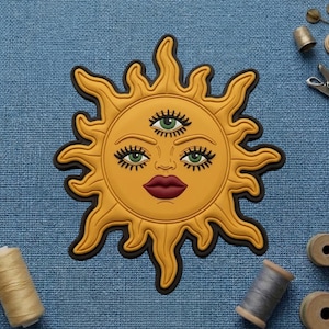 May include: A yellow sun-shaped patch with a face, three eyes, and a black outline. The sun has a smiling face with green eyes, long eyelashes, and dark red lips. The sun's rays are wavy.