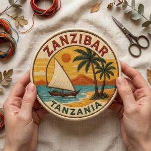 May include: A circular embroidery hoop featuring a stitched landscape of Zanzibar, Tanzania. The design depicts a dhow boat with a white sail on turquoise water, two palm trees on a sandy beach, and a sun against a yellow sky. The text ZANZIBAR TANZANIA is stitched in dark red and green.