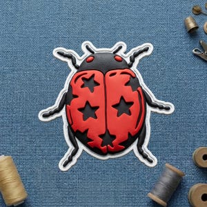 May include: A red and black ladybug-shaped patch with star cutouts on a blue textured surface. The ladybug has a white outline and is surrounded by sewing notions, including spools of thread and a thimble.