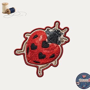 May include: An embroidered patch shaped like a ladybug with a red heart-shaped body featuring black heart spots and a black head. Two wooden spools of thread, one beige and one dark blue, with a needle and loose thread are positioned above.