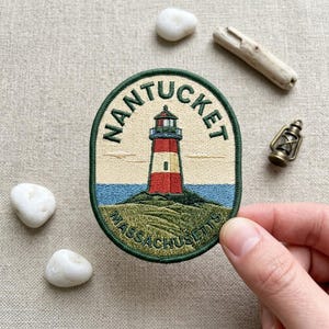 May include: An oval embroidered patch featuring a lighthouse with a red and white tower on a grassy hill. The text NANTUCKET is arched above the lighthouse, and MASSACHUSETTS is written below. The design includes a blue sky and sea background.