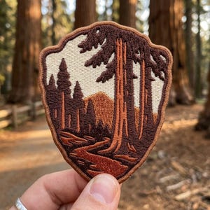May include: A shield-shaped embroidered patch featuring a landscape scene with tall evergreen trees, a winding path, and a mountain peak in the background. The design uses a color palette of cream, dark brown, and terracotta orange thread.