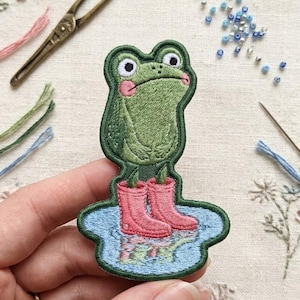 May include: An embroidered patch featuring a green frog with pink cheeks wearing bright pink rain boots while standing in a small blue puddle. The frog has large white eyes with black pupils and a neutral expression, rendered in detailed thread work.