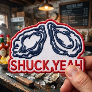 May include: An embroidered patch featuring two dark blue oyster illustrations outlined in red on a white background. The text SHUCK YEAH is embroidered in red capital letters at the bottom of the patch. The patch has a scalloped red border.