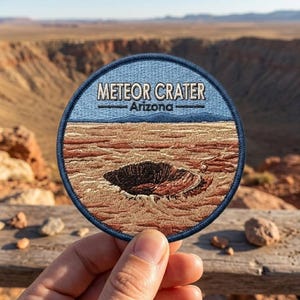May include: A circular embroidered patch featuring a landscape of the Meteor Crater in Arizona. The design includes a dark blue border, a light blue sky, and a tan and brown desert floor with a deep, dark brown impact crater in the center.